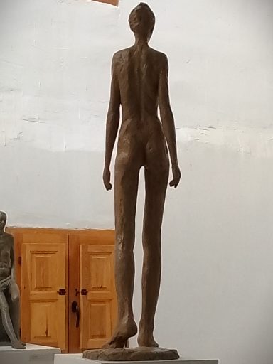 Exposición de escultura en el nuevo taller Escultura de una figura humana delgada, vista desde atrás, en un ambiente artístico.
