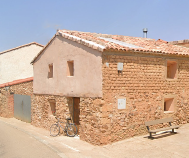 Nuevo taller en Gallocanta Casa de piedra con techo de tejas, bicicleta apoyada y banco en la calle.