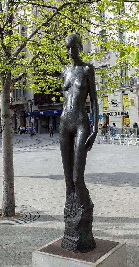 Escultura de mujer desnuda de bronce, rodeada de árboles y una calle concurrida.