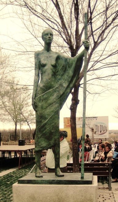 Estatua de bronce de una figura humana sosteniendo un bastón, rodeada de árboles.