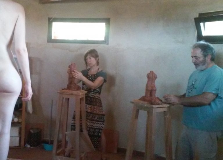 Clase de escultura con dos artistas trabajando en figuras de barro. La modelo, de espaldas en primer plano