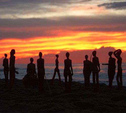 Siluetas de un conjunto de esculturas frente a un atardecer en la playa.