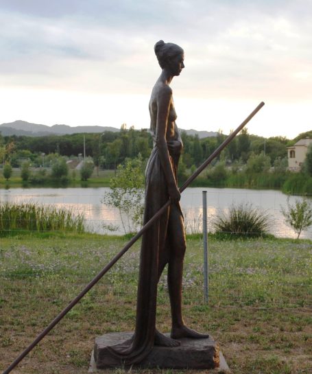 Escultura de mujer de perfil, sosteniendo una vara, con paisaje natural al fondo.