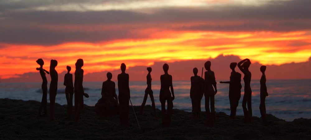 Siluetas de figuras humanas en la playa al atardecer, con cielo anaranjado y mar de fondo.