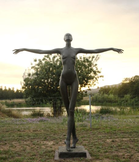 Escultura de una figura femenina con brazos extendidos frente a un paisaje natural.