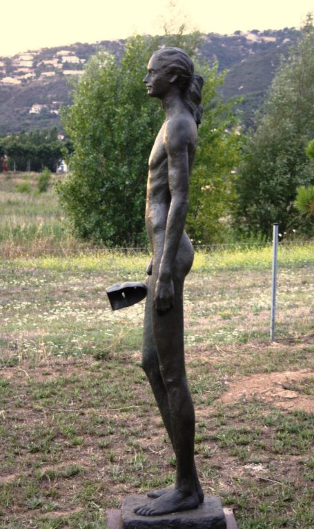 Escultura de una figura humana masculina de perfil, de pie en un paisaje natural. Sujeta una máscara con su mano derecha