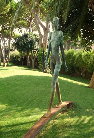 Escultura de una figura femenina de pie en un entorno verde y soleado.