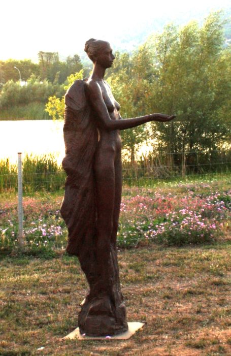 Escultura de mujer de pie, con dos grandes alas que recuerdan a la figura de un ángel, las manos extendidas, rodeada de flores y vegetación.