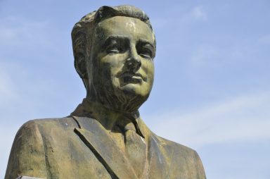 Estatua de un hombre con traje, mirada seria y fondo de cielo azul.