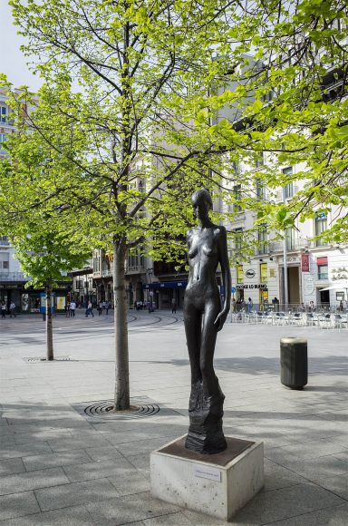 Escultura de bronce de figura femenina junto a árboles en una plaza urbana.