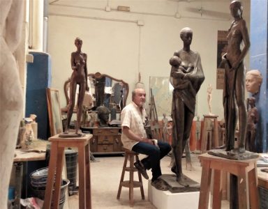 Escultura en un taller con un artista sentado entre estatuas de bronce.