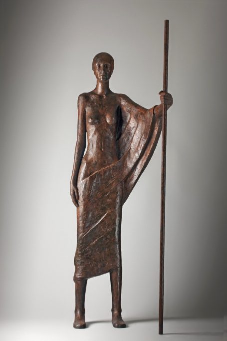 Escultura de mujer de bronce sosteniendo una vara, vestida con una falda drapeada.