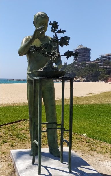 Escultura de un hombre cuidando un bonsái en una mesa, frente a la playa.