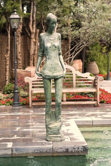 Escultura de bronce de figura femenina en un jardín con flores y una banca de madera.