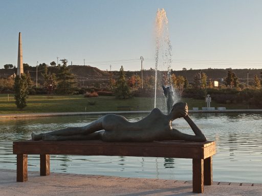 Escultura de bronce de una figura reclinada junto a un estanque y fuentes en un parque.