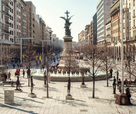 Plaza con fuente central y monumento, rodeada de calles y edificios urbanos.