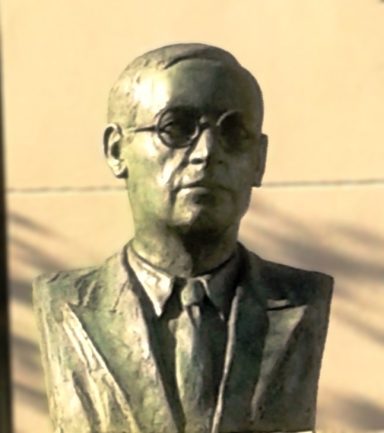 Busto de un hombre con gafas, vestido con traje, en un fondo claro.