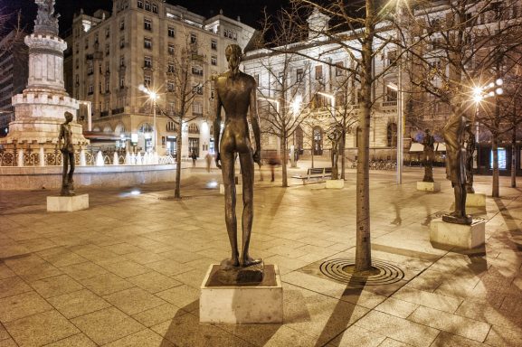 Estatuas en una plaza iluminada por la noche, rodeadas de árboles y edificios.