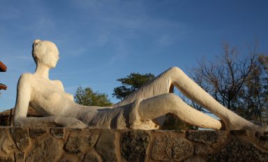 Escultura de una figura femenina tumbada, sobre una base de piedra, bajo un cielo azul.