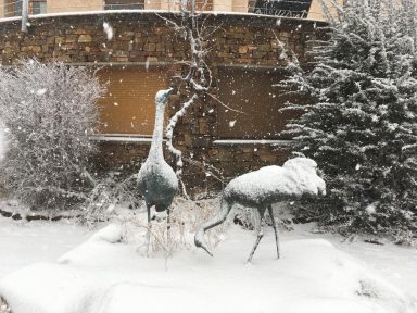 Escultura de dos grullas de bronce en un paisaje nevado con arbustos cubiertos de nieve.