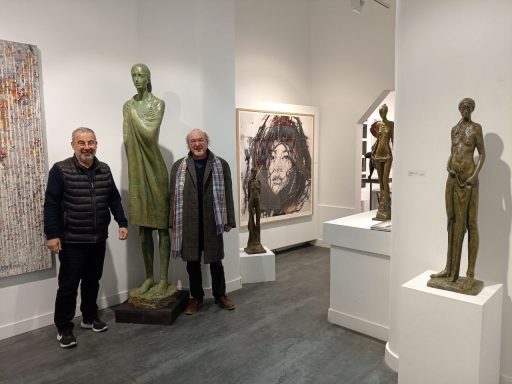 Dos hombres posando junto a una escultura en una galería de arte con distintas esculturas y pinturas