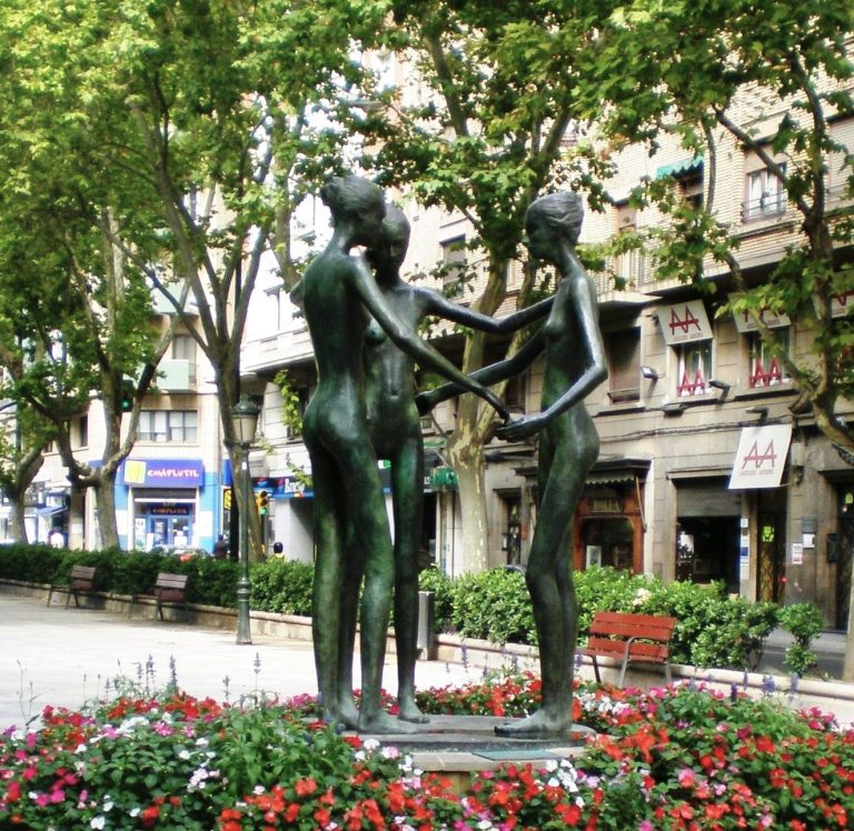 Escultura de tres figuras humanas unidas en un paseo urbano con árboles y flores.