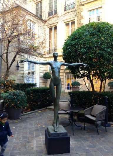 Escultura de figura humana de pie con brazos extendidos en un jardín urbano.