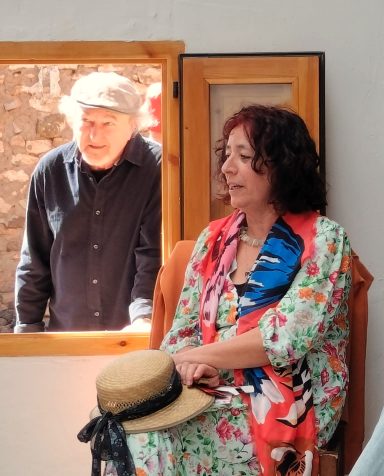 Una mujer sentada con un vestido florido hablando junto a un hombre que observa de buen humor desde la ventana que tiene junto a ella