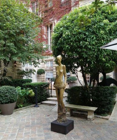 Escultura con un tono dorado de figura femenina en un jardín con arbustos y bancos.