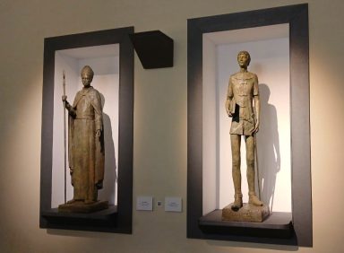 Estatuas de figuras humanas en vitrinas, una con un bastón y otra con un libro y una espada enfundada