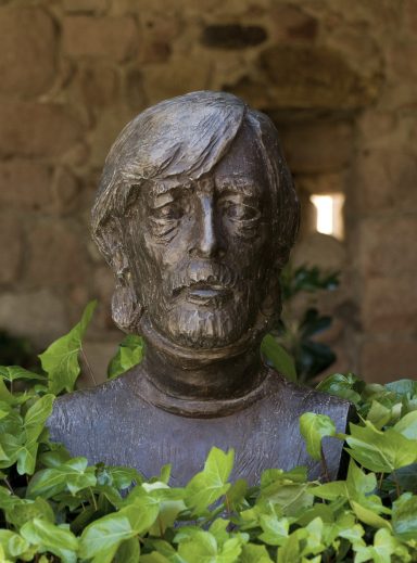 Busto de bronce de un hombre con cabello largo y barba, rodeado de plantas verdes.