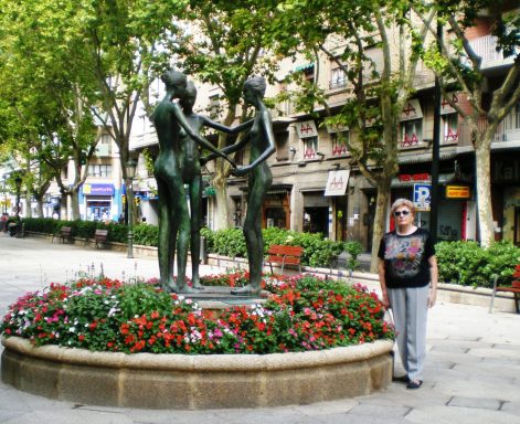 Una mujer posando junto a una escultura de tres mujeres en un paseo urbano con vegetación