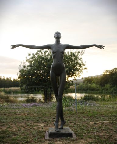 Escultura de figura humana con los brazos extendidos, frente a un paisaje natural.