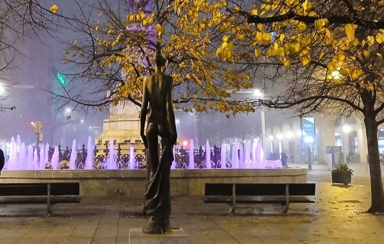 Estatua de figura femenina junto a fuente iluminada en un entorno urbano nocturno.