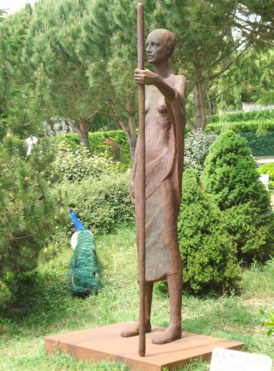 Escultura de figura humana, de pie con un bastón, en un entorno ajardinado.