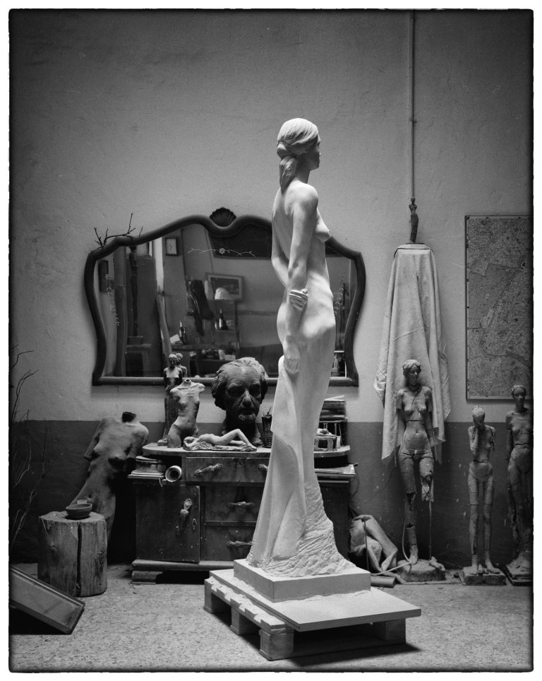 Escultura de una figura femenina en un taller, con un espejo y herramientas alrededor.