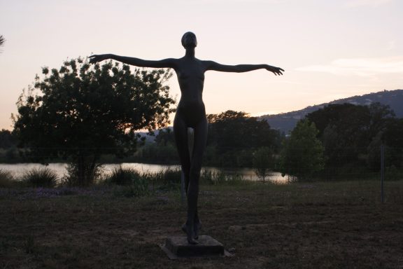 Escultura de figura humana con brazos extendidos, al atardecer junto a un lago.