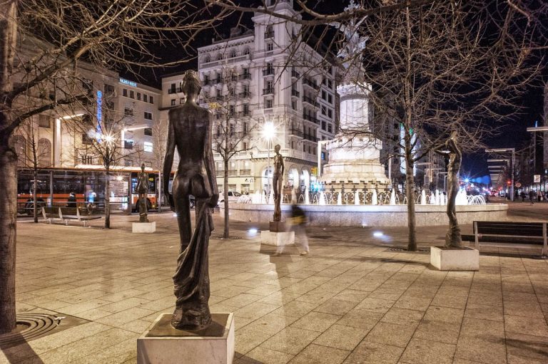 Estatua de mujer en una plaza iluminada, con fuente y edificios al fondo por la noche.