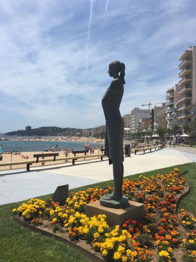 Escultura estilizada frente a un mar y una playa, rodeada de flores.