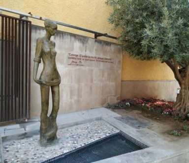 Escultura de mujer junto a un estanque y pared con inscripción en un entorno ajardinado.