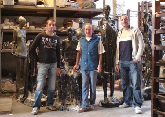 Tres hombres junto a esculturas en un taller de fundición artística.