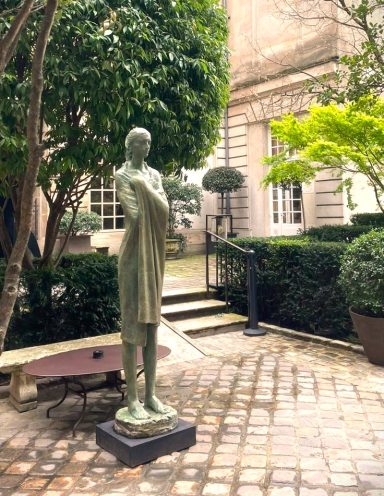 Escultura de una figura femenina de pie en un jardín con plantas y una terraza.