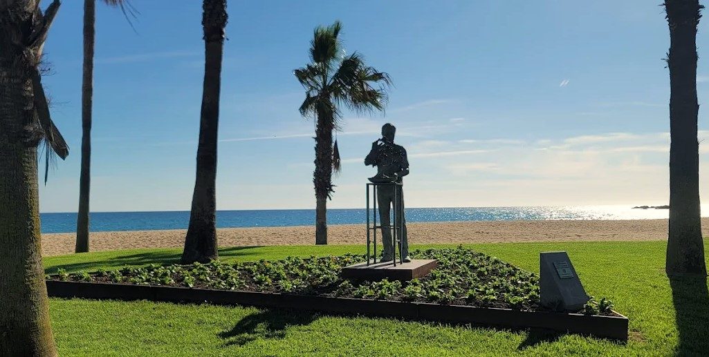 Estatua de un hombre en una playa, rodeada de palmeras y césped verde.