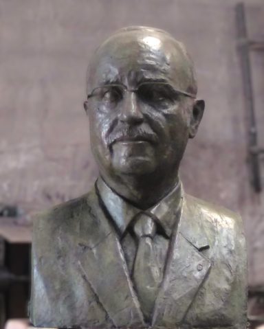 Busto de bronce de un hombre con traje y corbata, expresión seria.